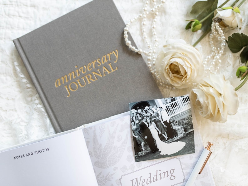 May include: A gray linen-covered journal with the words "anniversary journal" embossed in gold. The journal is open to a blank page with the words "notes and photos" printed at the top. A black and white photo of a wedding party is on the right page with the word "wedding" printed below it. A string of pearls and white flowers are on the top of the journal.