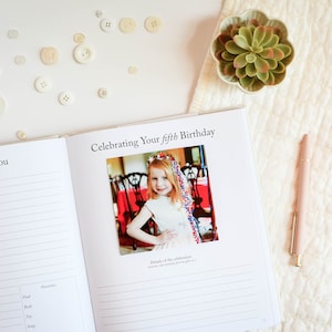 May include: A white book with the title "Celebrating Your Fifth Birthday" and a photo of a young girl in a white dress with a red, white and blue headband. The book has a section titled "All About You" with lines for writing.