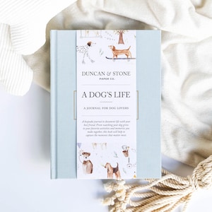 May include: A light blue journal titled "A Dog's Life" with a decorative paper band featuring dog illustrations. The journal is for dog lovers and includes the text "A journal for dog lovers". The journal is surrounded by a white plush toy and a cream-colored blanket.