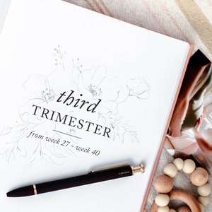 May include: A white journal with a floral design and the text "third trimester from week 27 - week 40". A black pen and a wooden teething ring are on the page.
