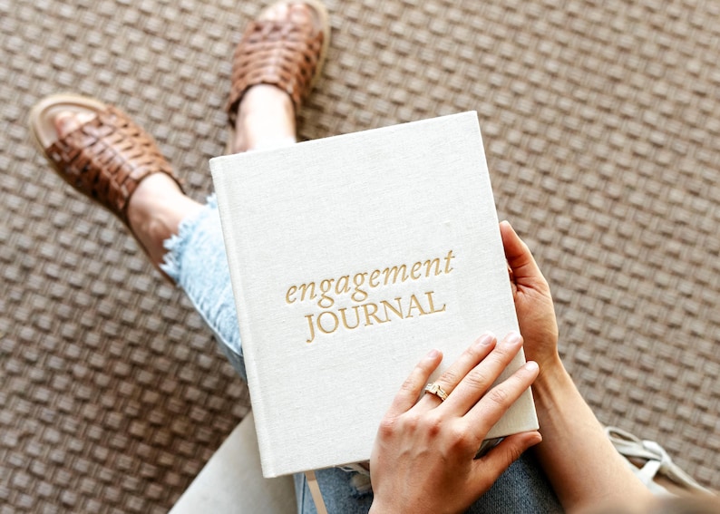 Peut inclure: Un journal blanc recouvert de lin avec le texte dor&eacute; "engagement JOURNAL" sur la couverture. Le journal est tenu par une personne portant un jean bleu.