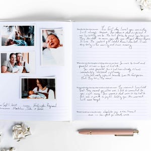 May include: Open journal with photos of a newborn baby and handwritten notes. The journal includes photos of the baby with family members. A pink pen rests on the white surface. White flowers are scattered around the journal.