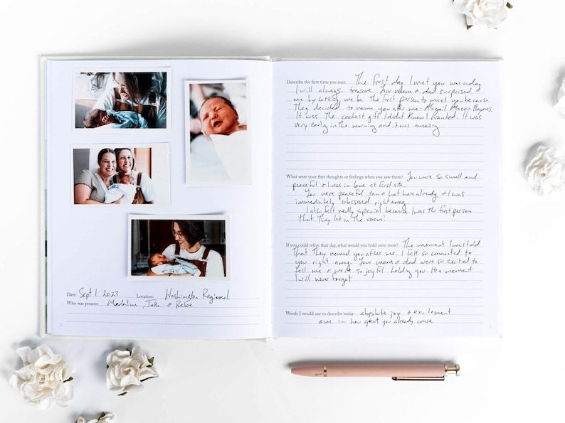 May include: Open journal with photos of a newborn baby and handwritten notes. The journal includes photos of the baby with family members. A pink pen rests on the white surface. White flowers are scattered around the journal.