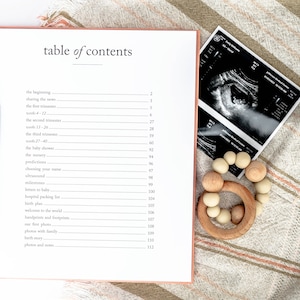 May include: An open book with a table of contents for a pregnancy journal. The book is open to the page with the table of contents. The book is on a light brown and white striped blanket. There is a pen, a wooden baby teether, and a black and white ultrasound image on the blanket.