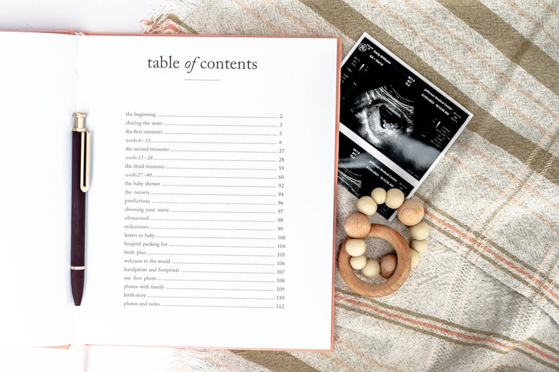 May include: An open book with a table of contents for a pregnancy journal. The book is open to the page with the table of contents. The book is on a light brown and white striped blanket. There is a pen, a wooden baby teether, and a black and white ultrasound image on the blanket.