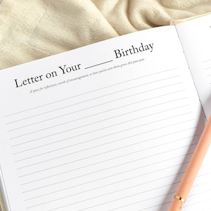 May include: Open journal with lined pages and the text "Letter on Your Birthday." A peach-colored pen with gold accents rests on the right page. The journal is open on a light-colored surface.