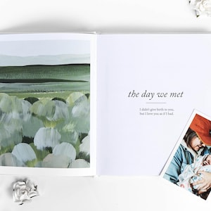 May include: An open book with the text "the day we met" and a photo of a person holding a newborn baby. The left page features a green and white abstract landscape painting. White flowers are scattered around the book.