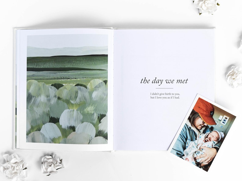 May include: An open book with the text "the day we met" and a photo of a person holding a newborn baby. The left page features a green and white abstract landscape painting. White flowers are scattered around the book.