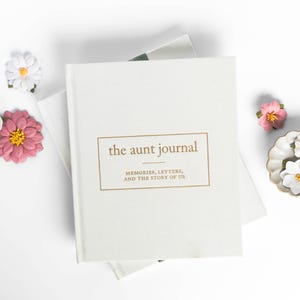 May include: A cream-colored journal titled "the aunt journal" with gold lettering, surrounded by paper flowers in pink, white, and yellow. The journal's cover reads "MEMORIES, LETTERS, AND THE STORY OF US."