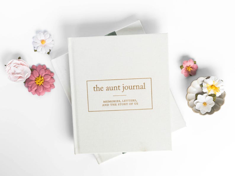 May include: A cream-colored journal titled "the aunt journal" with gold lettering, surrounded by paper flowers in pink, white, and yellow. The journal's cover reads "MEMORIES, LETTERS, AND THE STORY OF US."