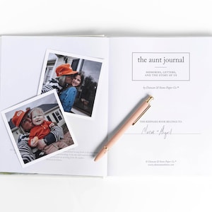 May include: An open book with the title "the aunt journal" and the subtitle "MEMORIES, LETTERS, AND THE STORY OF US." Two photos are placed on the left page. A pink pen rests on the right page. The book is white.