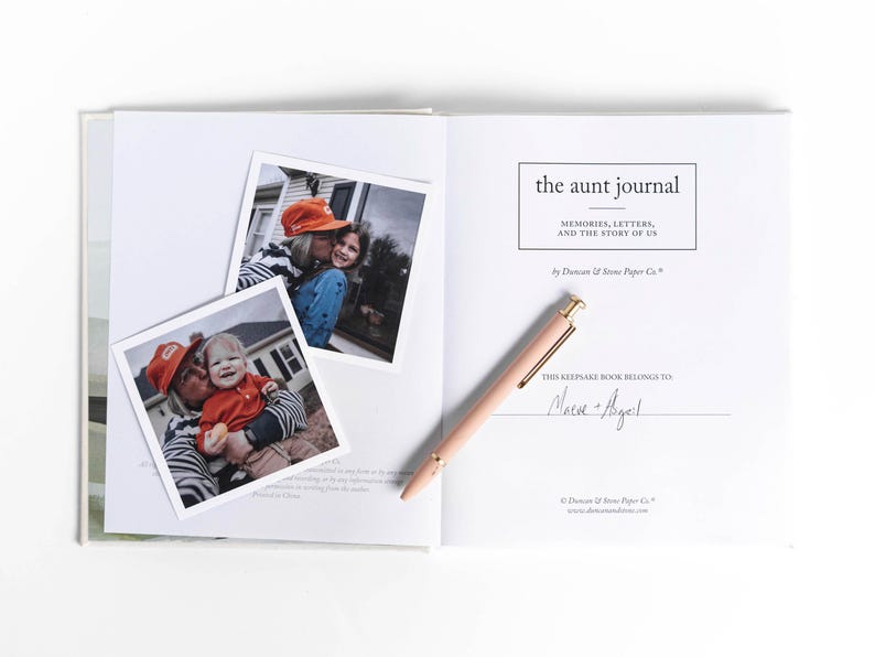 May include: An open book with the title "the aunt journal" and the subtitle "MEMORIES, LETTERS, AND THE STORY OF US." Two photos are placed on the left page. A pink pen rests on the right page. The book is white.