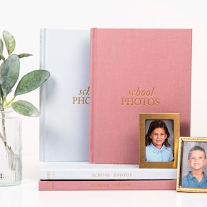 K&ouml;nnte beinhalten: Zwei rosa und blaue Fotoalben mit dem Text "school photos" auf der Vorderseite. Die Alben sind &uuml;bereinander gestapelt. Es gibt zwei kleine goldene Bilderrahmen mit Fotos von Kindern.