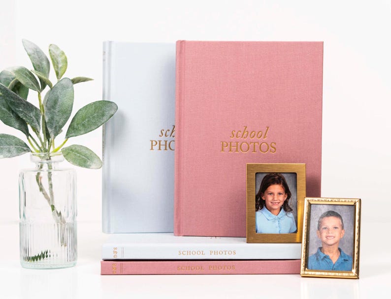 K&ouml;nnte beinhalten: Zwei rosa und blaue Fotoalben mit dem Text "school photos" auf der Vorderseite. Die Alben sind &uuml;bereinander gestapelt. Es gibt zwei kleine goldene Bilderrahmen mit Fotos von Kindern.
