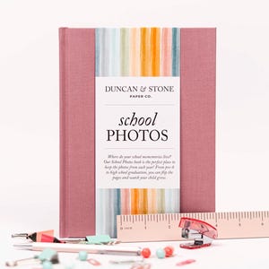 May include: A pink photo album with a colorful striped design on the front cover. The album is titled "School Photos" and has the text "Duncan & Stone Paper Co." on the front cover.