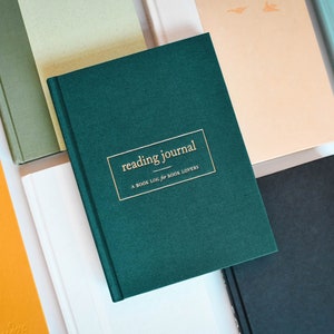 May include: A green hardcover journal with gold lettering that reads "reading journal, a book log for book lovers". The journal is surrounded by other books in various colors and textures.