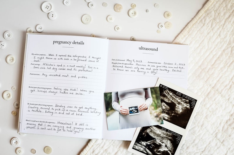 May include: A white journal with a black cover and the words "pregnancy details" and "ultrasound" written on the front. The journal is open to two pages, one with handwritten notes about pregnancy cravings and the other with a photo of a pregnant woman holding an ultrasound image and two ultrasound images.