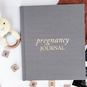 May include: A gray pregnancy journal with gold lettering that reads "pregnancy journal". The journal is surrounded by baby-related items, including a crocheted bunny rattle, wooden letter blocks, and ultrasound images.