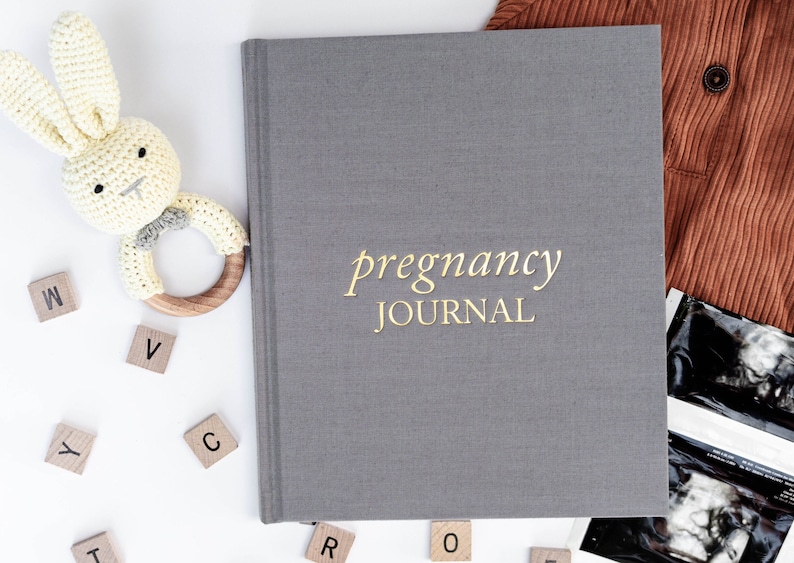 May include: A gray pregnancy journal with gold lettering that reads "pregnancy journal". The journal is surrounded by baby-related items, including a crocheted bunny rattle, wooden letter blocks, and ultrasound images.