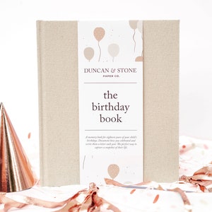 May include: A beige linen-covered journal with the title "The Birthday Book" and the text "Duncan & Stone Paper Co." on the front cover. The cover features a design of balloons in pink and beige.