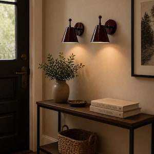 May include: Two burgundy wall sconces with conical shades light a hallway. A wooden console table holds a woven basket, books, and a vase with greenery. The books read "Live Beautifully" and "This is Home."