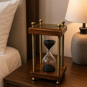 May include: A rectangular hourglass featuring a dark wood frame with four brass-toned metal pillars at the corners. The glass bulb contains black sand. The top and bottom plates are made of wood with brass-toned metal accents and small spherical feet.