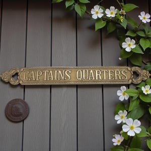 Puede incluir: Un letrero metálico dorado que dice "CAPTAINS QUARTERS" en mayúsculas. El letrero tiene adornos decorativos en cada extremo y está montado sobre una superficie de madera gris. Una pequeña placa redonda de color bronce está debajo del letrero. Flores blancas y hojas verdes en el fondo.