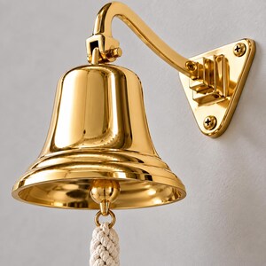 May include: A polished gold-tone wall-mounted bell featuring a smooth, reflective surface and a curved metal arm attached to a triangular base with three visible screws. A thick, braided off-white rope pull hangs from the center clapper.