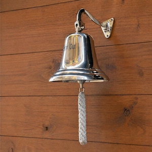 May include: A polished silver bell with a curved arm and mounting plate, attached to a wooden wall. The bell has a rope pull and the words "Hon' Out" are etched on the front. The bell is a nautical or maritime themed item.