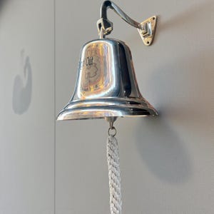 May include: A polished silver bell with the word "Out" visible on the front. The bell is mounted on a wall with a curved arm and a braided rope pull. The bell is a nautical or maritime-themed item.