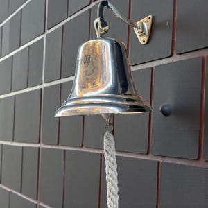 May include: A polished silver bell with a rope pull, mounted on a dark wall. The bell has the word "Out" visible on its surface. The bell is attached to a curved arm and a wall mount.