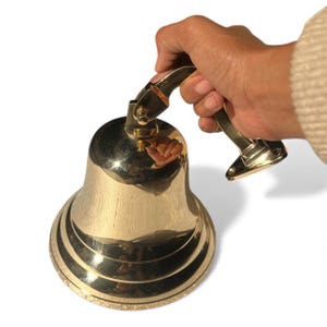 May include: A shiny brass bell with a curved handle. The bell has a flared base with concentric rings and a polished surface. The handle is attached to the top of the bell. The bell is held by a hand.