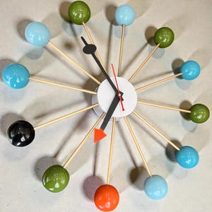 Może przedstawiać: Zegar ścienny w stylu retro z białą tarczą, czarnymi wskazówkami i czerwoną wskazówką sekundową. Posiada kolorowe kule w kolorze niebieskim, zielonym, czarnym i pomarańczowym, przymocowane do drewnianych prętów. Unikalny element wystroju wnętrz.