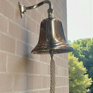 May include: A polished brass bell mounted on a brick wall. The bell has a curved arm and a rope pull. The bell is a shiny gold color and reflects the surrounding environment. The bell is likely used for signaling or decoration.