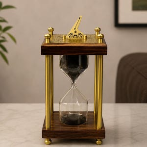 Puede incluir: Un reloj de arena rectangular con marco de madera oscura, cuatro pilares de latón pulido y pies esféricos. La placa superior de latón presenta grabados y un gnomon triangular central. El bulbo de cristal contiene arena negra fina.