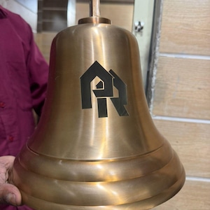 May include: A large, bronze-colored bell with a handle and a logo. The bell has a smooth, polished surface and a flared base. The logo is black and features a house silhouette and the letters "PR".