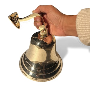 May include: A polished brass bell with a curved bracket for mounting. The bell has a flared base with concentric rings and a clapper inside. The bracket has a mounting plate with screw holes. The bell is held by a hand.