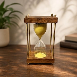 Könnte beinhalten: Eine Sanduhr mit einem dunkelbraunen Holzrahmen, vier vertikalen Messingstäben und einem klaren Glaskolben, der hellgelben Sand enthält. Die oberen und unteren Holzteile sind quadratisch und an jeder Ecke mit kleinen Messingkugeln verziert.