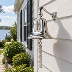 May include: A polished silver bell with the text "Hoin' Out" is mounted on a white exterior wall. The bell is attached to a curved arm and has a rope pull. The background shows a house, greenery, and a body of water.