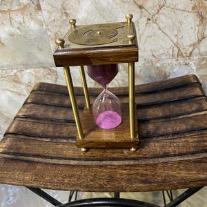 May include: A square hourglass featuring a dark wood base and top, four brass pillars, and a glass bulb filled with vibrant pink sand. The top brass plate displays an engraved nautical compass design with an anchor symbol and directional markings.