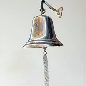 May include: A polished chrome bell with a curved arm and mounting plate. The bell has the words "Keep Out" visible on its surface. A thick, gray rope hangs from the bell's clapper. The bell is designed for nautical or decorative use.