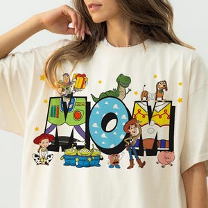 May include: Cream-colored oversized t-shirt featuring the word "MOM" in colorful block letters with Toy Story characters. The "O" is blue with cloud details, and stars are scattered around the letters. The shirt has short sleeves.