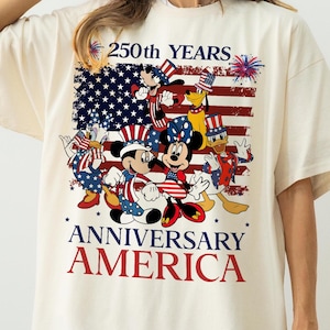 May include: Cream-colored t-shirt featuring a graphic of Mickey Mouse, Minnie Mouse, Donald Duck, and Goofy in patriotic attire. The design includes an American flag and the text "250th YEARS ANNIVERSARY AMERICA" in red and blue.