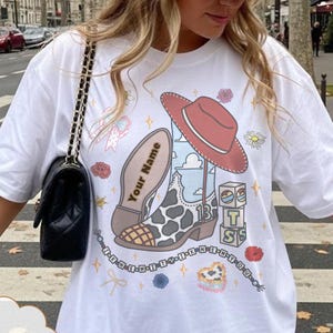 Op de afbeelding: Een wit t-shirt met een grafische print van een cowboylaars met koeienprint, een rode cowboyhoed, gestapelde blokken met de letters T, S, S en de tekst Your Name op de zool van de laars, inclusief kleine bloem- en steraccenten.