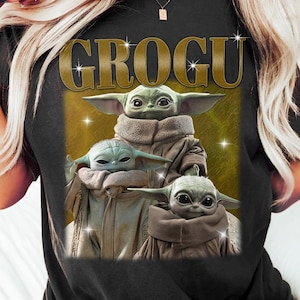 K&ouml;nnte beinhalten: Schwarzes T-Shirt mit dem Wort "GROGU" in goldfarbenen Buchstaben &uuml;ber drei Bildern von Grogu, auch bekannt als Baby Yoda, aus der Star Wars-Serie. Die Bilder zeigen Grogu in einem beigen Gewand mit gro&szlig;en Ohren und Glitzereffekten.