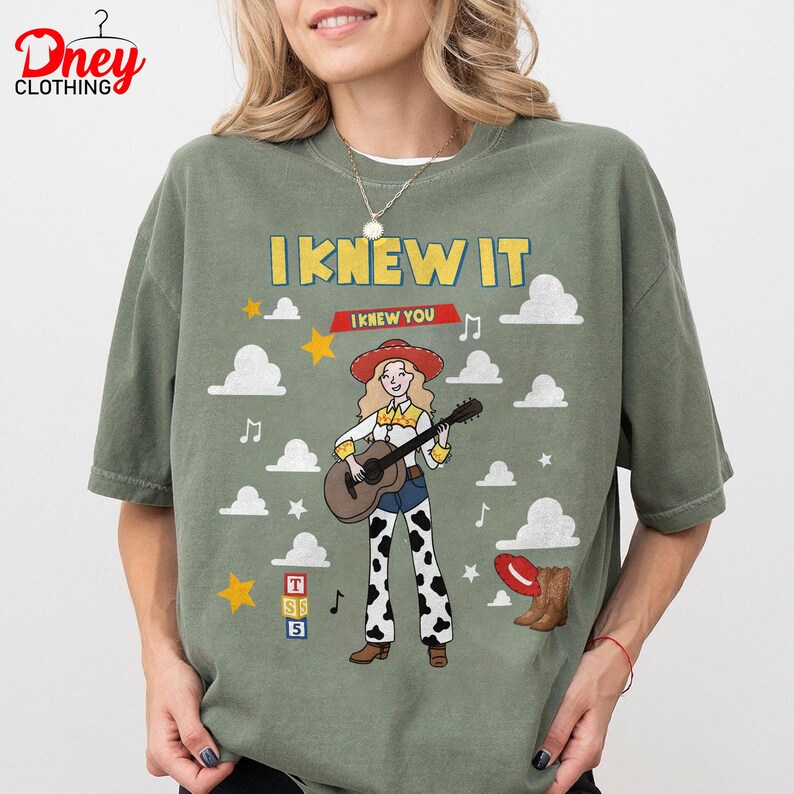 May include: An olive green t-shirt featuring a graphic of a blonde character in a red cowboy hat playing an acoustic guitar. The text reads I KNEW IT and I KNEW YOU. The design includes white clouds, yellow stars, musical notes, a red boot, and blocks.