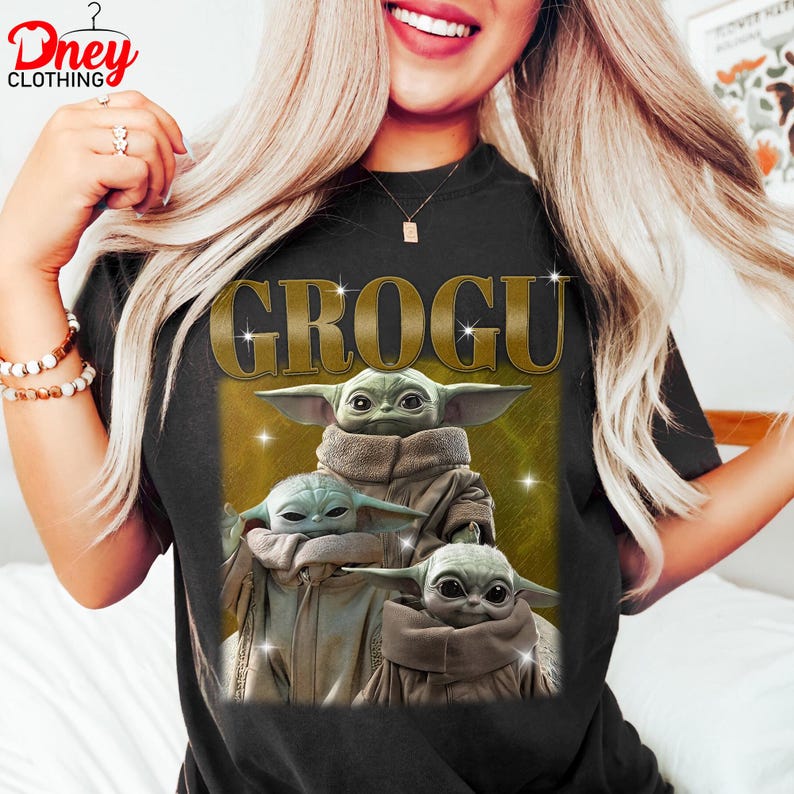 K&ouml;nnte beinhalten: Schwarzes T-Shirt mit dem Wort "GROGU" in goldfarbenen Buchstaben &uuml;ber drei Bildern von Grogu, auch bekannt als Baby Yoda, aus der Star Wars-Serie. Die Bilder zeigen Grogu in einem beigen Gewand mit gro&szlig;en Ohren und Glitzereffekten.