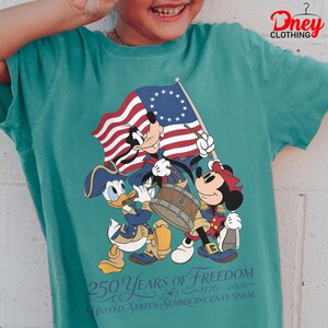 May include: Teal t-shirt with a patriotic Disney design. Goofy, Donald Duck, and Minnie Mouse are depicted in Revolutionary War outfits, holding a flag and playing instruments. The shirt displays the text "250 Years of Freedom 1776-2026 United States Semiquincentennial."