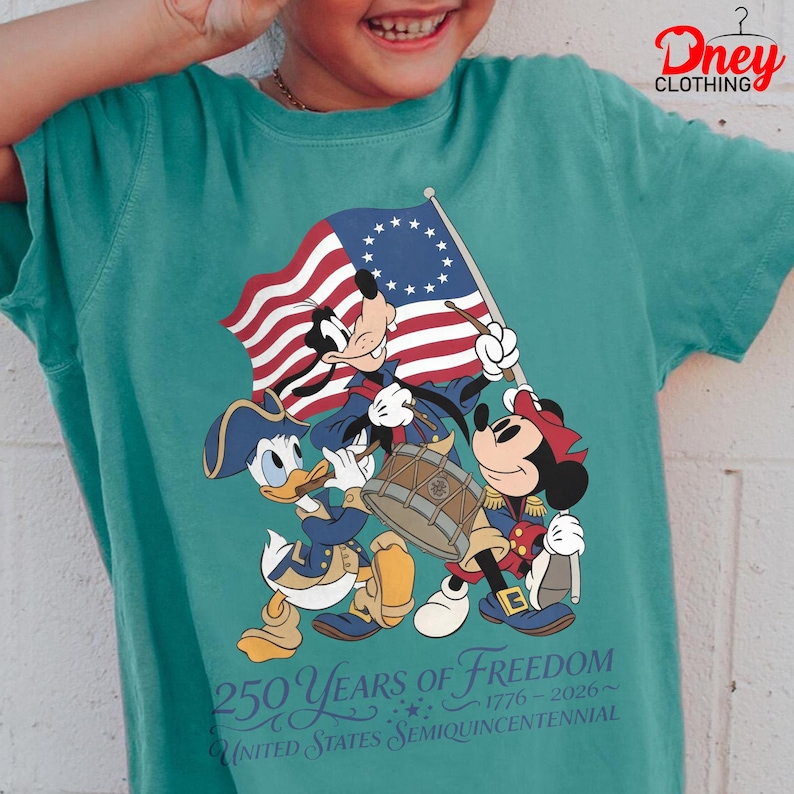 May include: Teal t-shirt with a patriotic Disney design. Goofy, Donald Duck, and Minnie Mouse are depicted in Revolutionary War outfits, holding a flag and playing instruments. The shirt displays the text "250 Years of Freedom 1776-2026 United States Semiquincentennial."