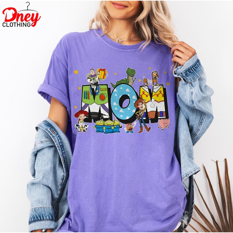 May include: A lilac t-shirt with the word "MOM" in colourful block letters, featuring characters from the Toy Story film. The shirt has short sleeves and a crew neck.